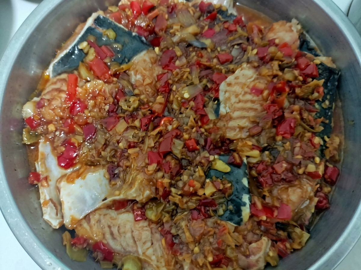 Un vassoio per la cottura al vapore in acciaio inossidabile con pezzi di pesce bianco ricoperti da una vivace salsa rossa di peperoncini tritati e aglio.