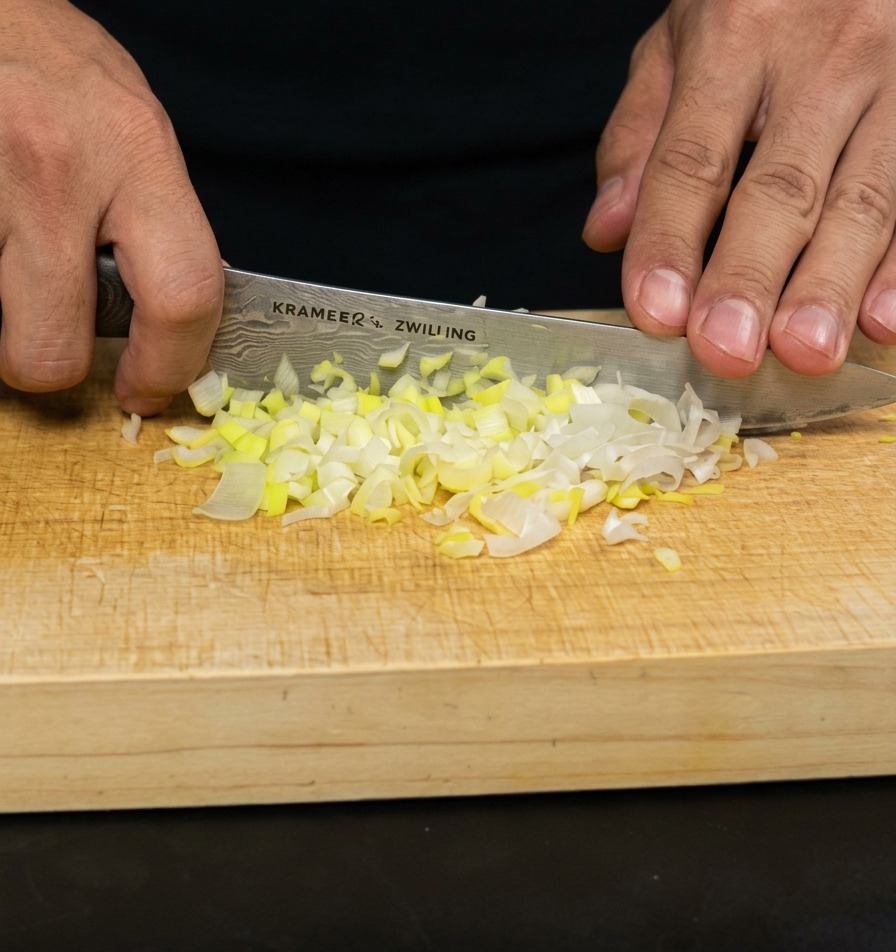 Mani che utilizzano un coltello da chef per tritare finemente dei cipollotti su un tagliere di legno chiaro.