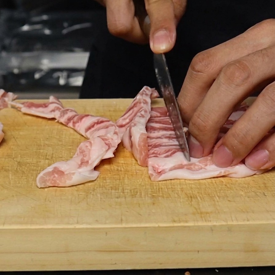Pancetta di maiale cruda tagliata in strisce sottili e uniformi di circa un centimetro su un tagliere di legno.