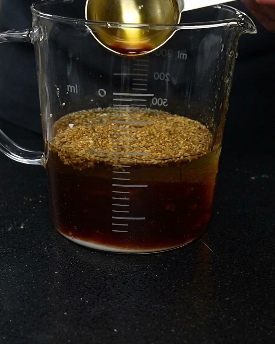 Un misurino dorato che versa salsa di soia scura in un contenitore di vetro con acqua e varie polveri di condimento.