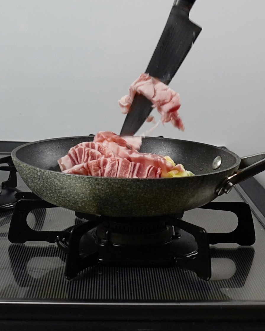 Un coltello che spinge le strisce di pancetta cruda in una padella contenente cipollotto tritato e aglio.