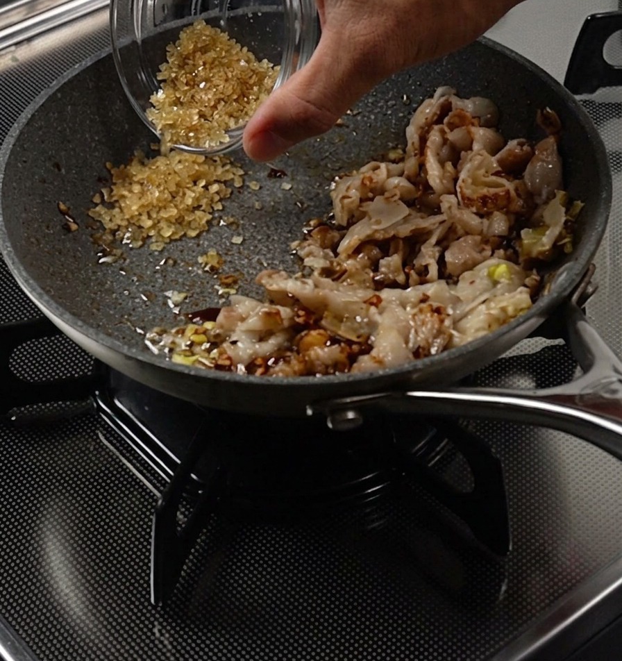 Una mano che versa zucchero da una ciotolina in unarea libera della padella accanto alla carne cotta.