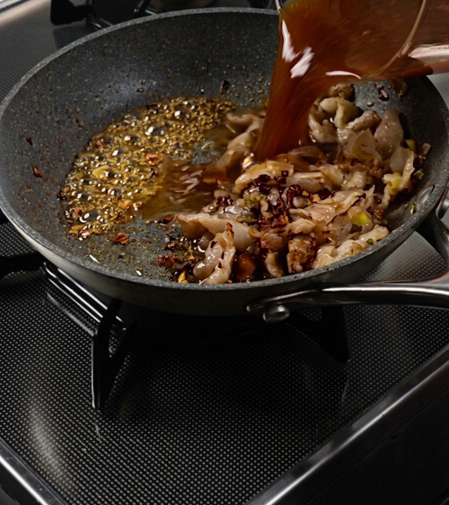 Salsa marrone versata da un misurino in una padella con pancetta e caramello che bolle.