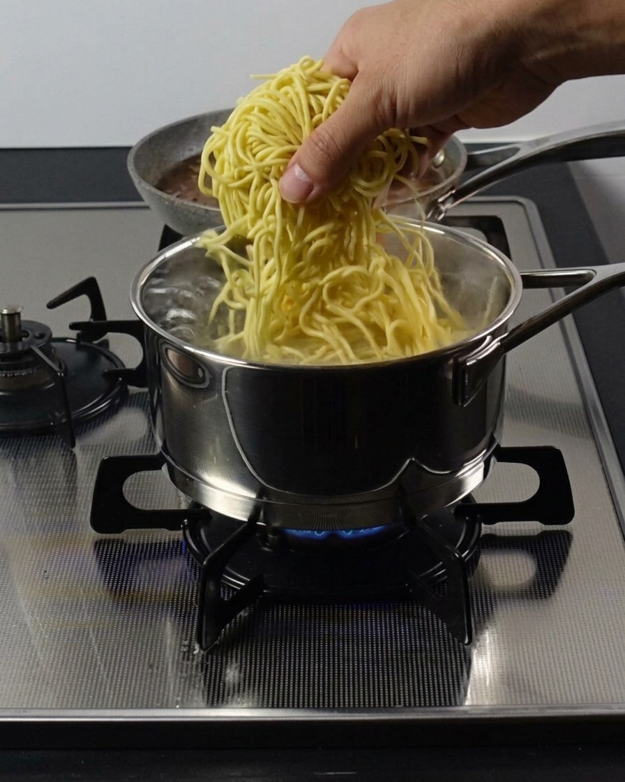 Una mano che immerge un nido di ramen gialli e spessi in una pentola di acciaio con acqua bollente.