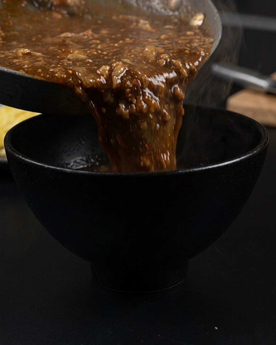 Brodo denso e scuro con pezzi di maiale versato da una padella in una ciotola nera opaca, con vapore che sale.