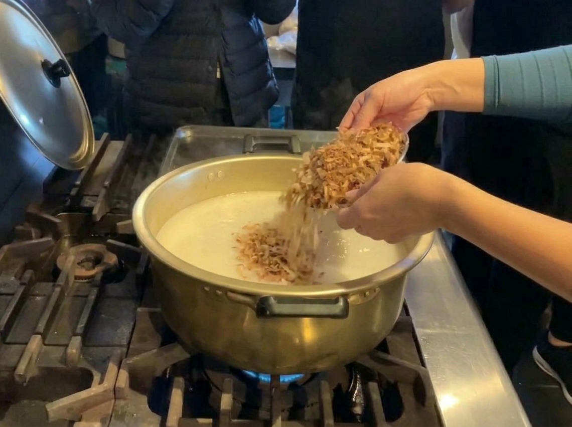 Una persona versa una grande quantità di scaglie di pesce essiccato da una ciotola dargento in una pentola dorata di brodo bianco.