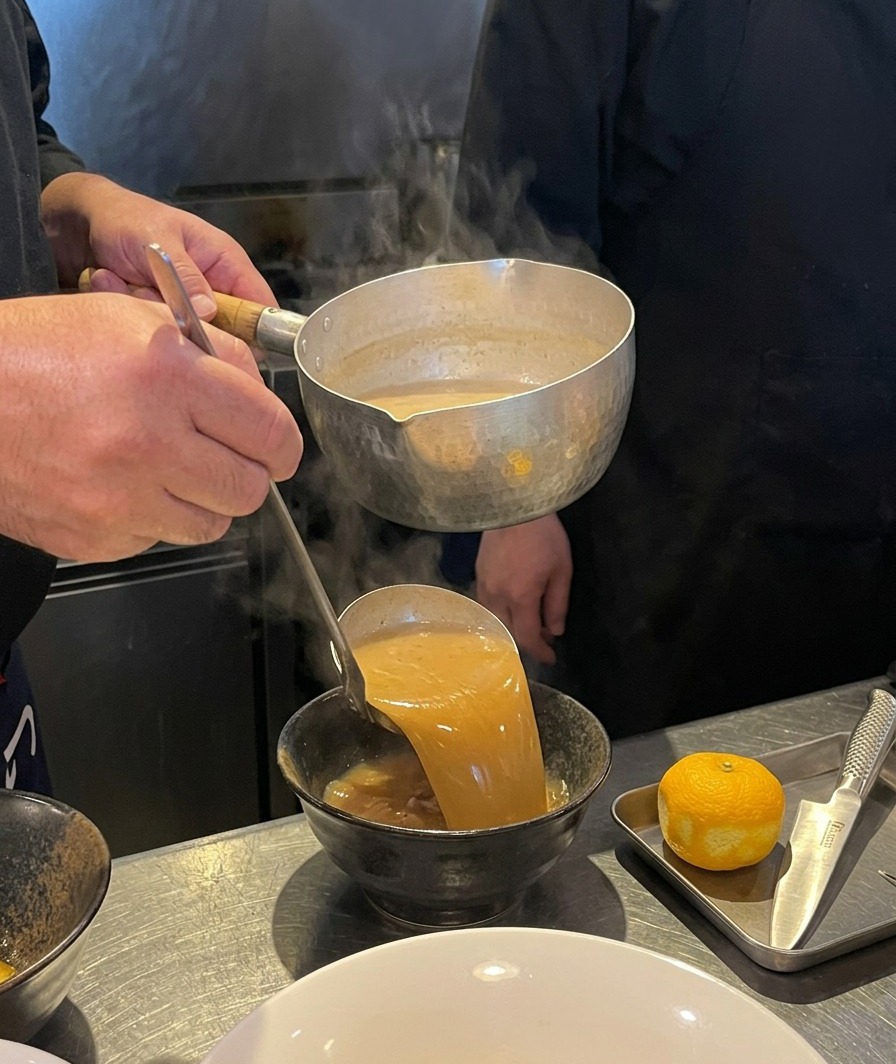 Brodo marrone denso che viene versato da un mestolo in una ciotola di ceramica nera.