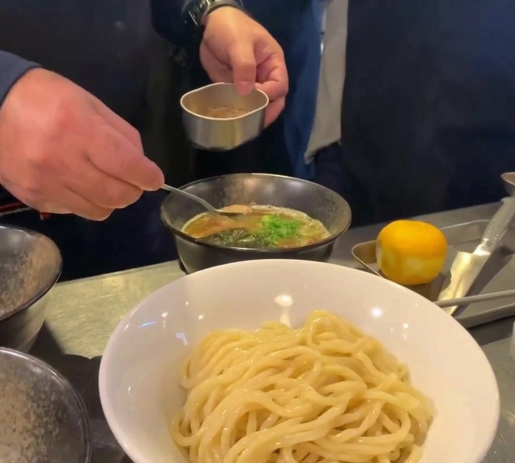 Un cucchiaino deposita una montagnola di polvere di pesce marrone in una ciotola di brodo scuro guarnito con cipollotti.
