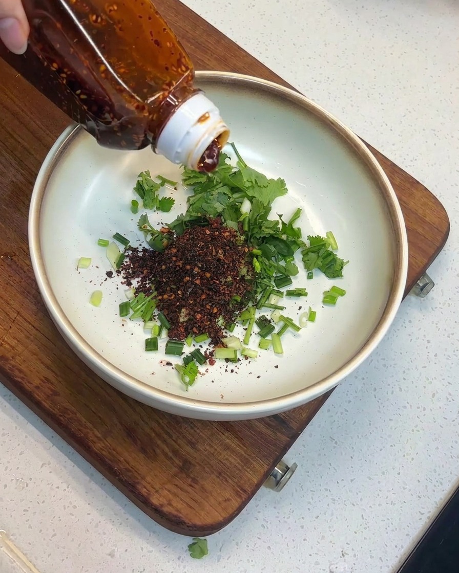 Olio di peperoncino rosso versato da una bottiglia su fiocchi di peperoncino e erbe in una ciotola.