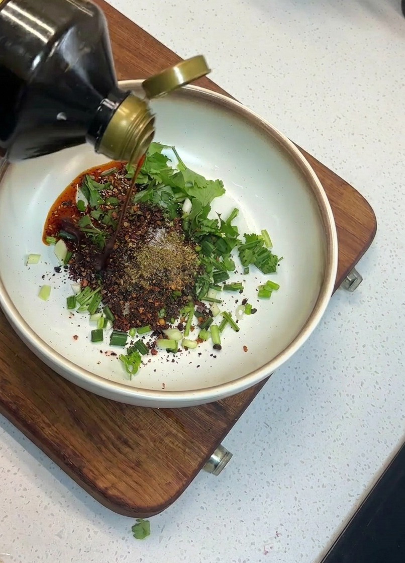 Una mano versa salsa di soia da una bottiglia di vetro in una ciotola con cipollotti, coriandolo e peperoncino.