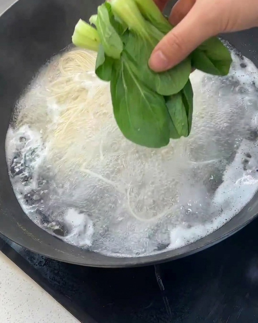Una mano aggiunge bok choy verde fresco in un wok con noodles in cottura.
