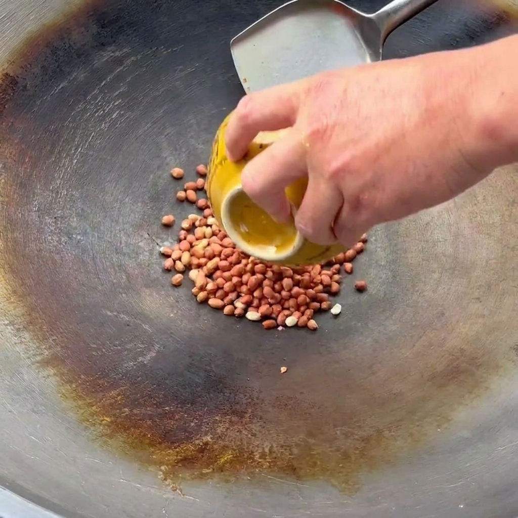 Una mano versa arachidi crude da una ciotola gialla in un wok per saltarle con una spatola.