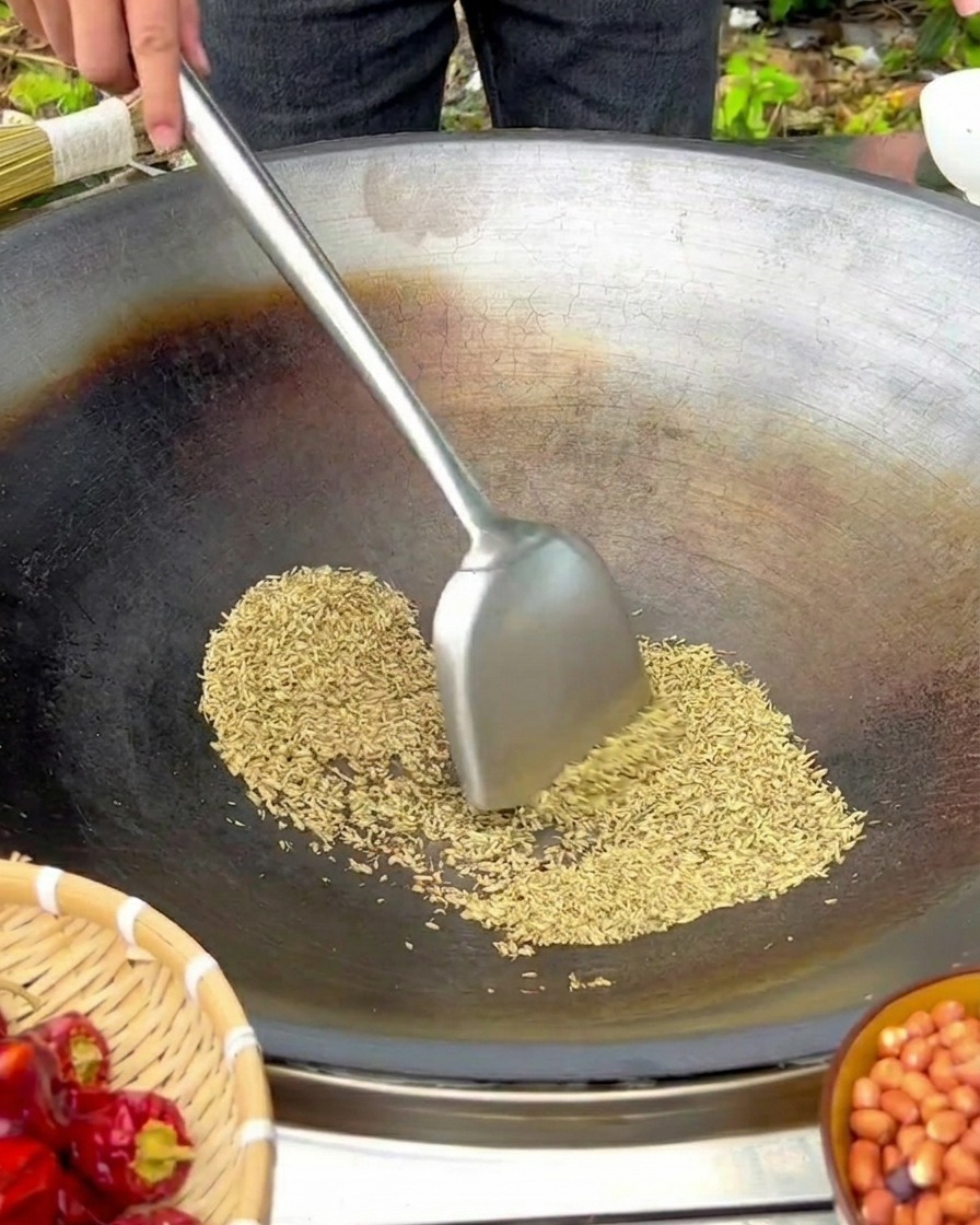 Semi di finocchio mescolati con una spatola in un grande wok di ferro a fuoco basso.