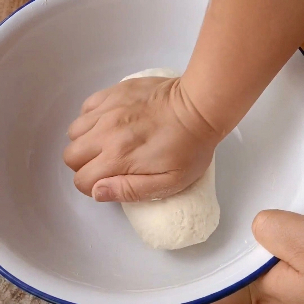 Due mani che impastano con decisione una palla di pasta allinterno di una ciotola bianca.
