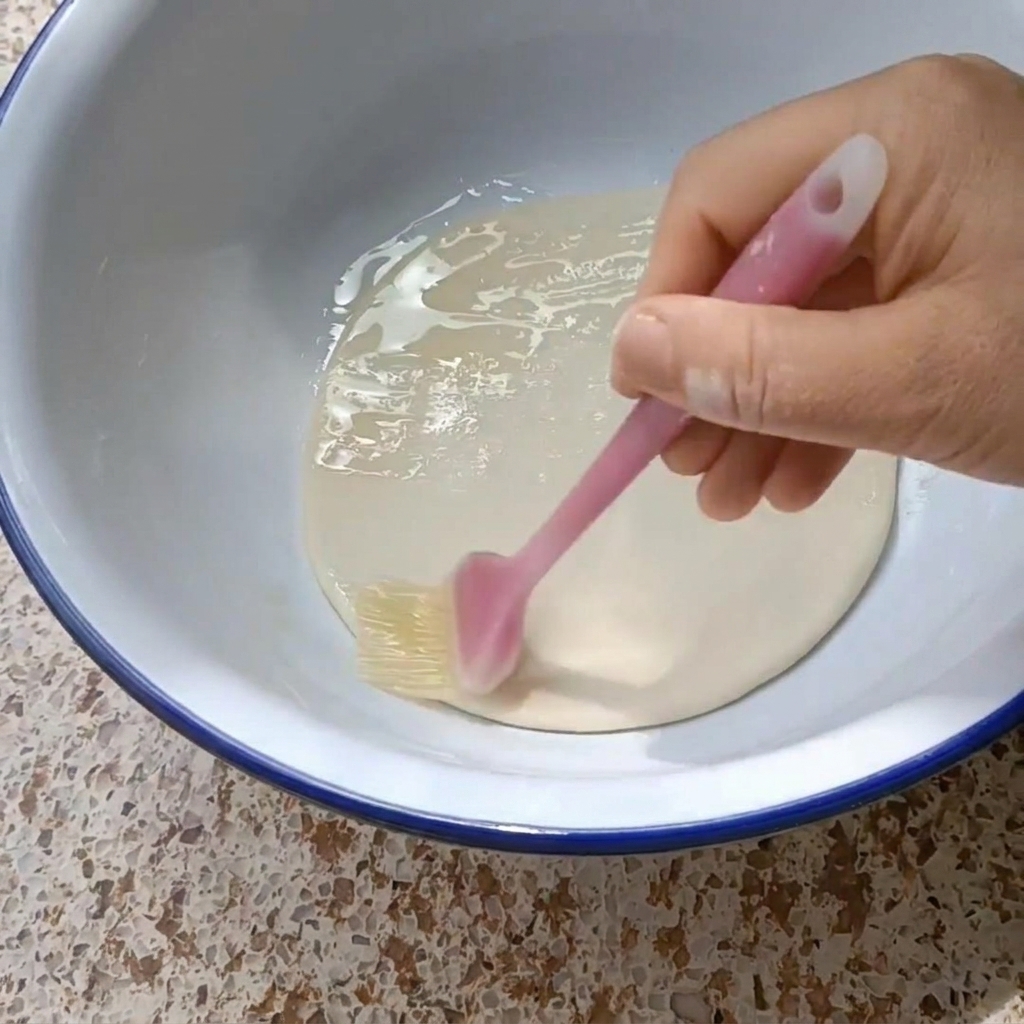 Una mano che usa un pennello in silicone per rivestire un disco di pasta con olio vegetale.