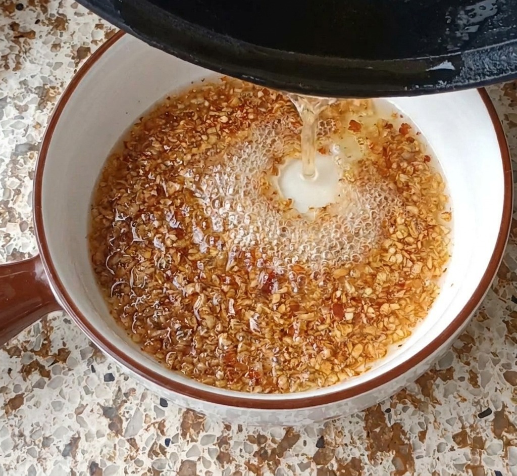 Olio bollente versato da una padella in una ciotola con aglio e peperoncino, creando bolle e fumo.