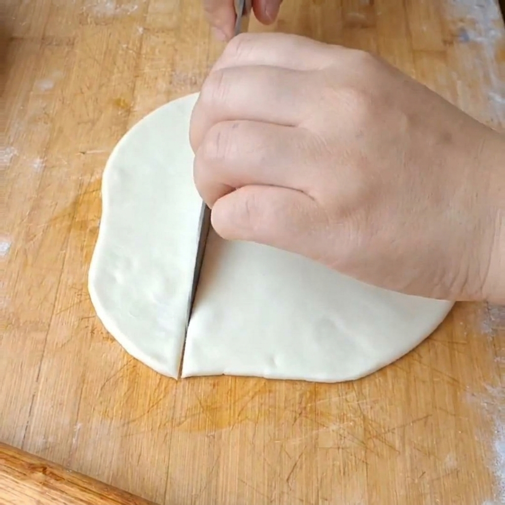 Una mano che usa un coltello grande per tagliare un disco di pasta piatto in larghe strisce su un tagliere.