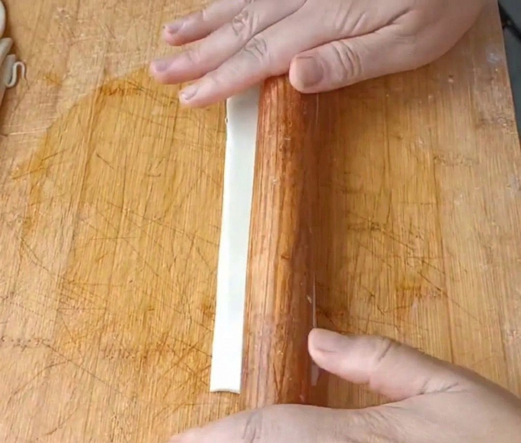 Primo piano di mani che usano un mattarello per appiattire una lunga striscia di pasta su un tagliere di legno.