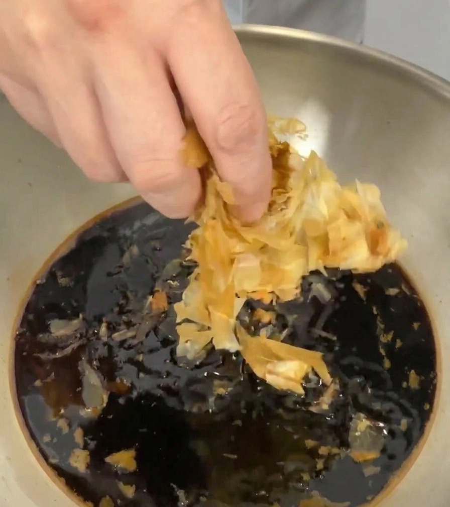Mano che immerge sottili fiocchi di katsuobushi nella salsa scura allinterno del pentolino.