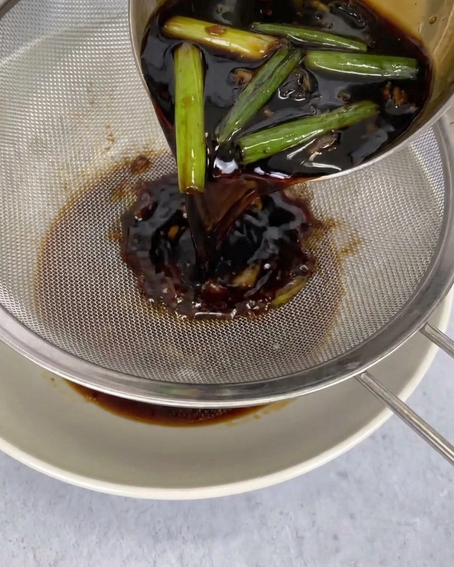Salsa scura versata attraverso un colino a maglie fini in una ciotola, trattenendo alga e peperoncini.