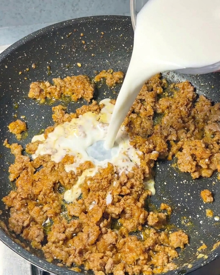 Un flusso di latte bianco versato da un contenitore in vetro in una padella con carne di maiale e condimenti.
