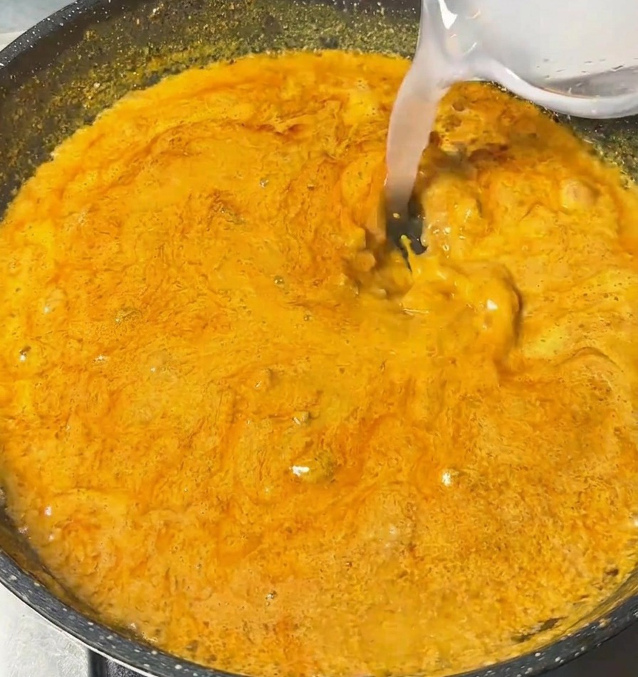Liquido trasparente versato da una ciotolina in una padella con salsa al curry arancione e densa.