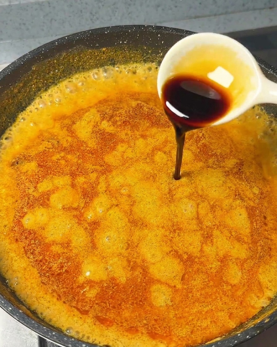 Un cucchiaio bianco che versa salsa di soia scura in una salsa al curry arancione in padella.