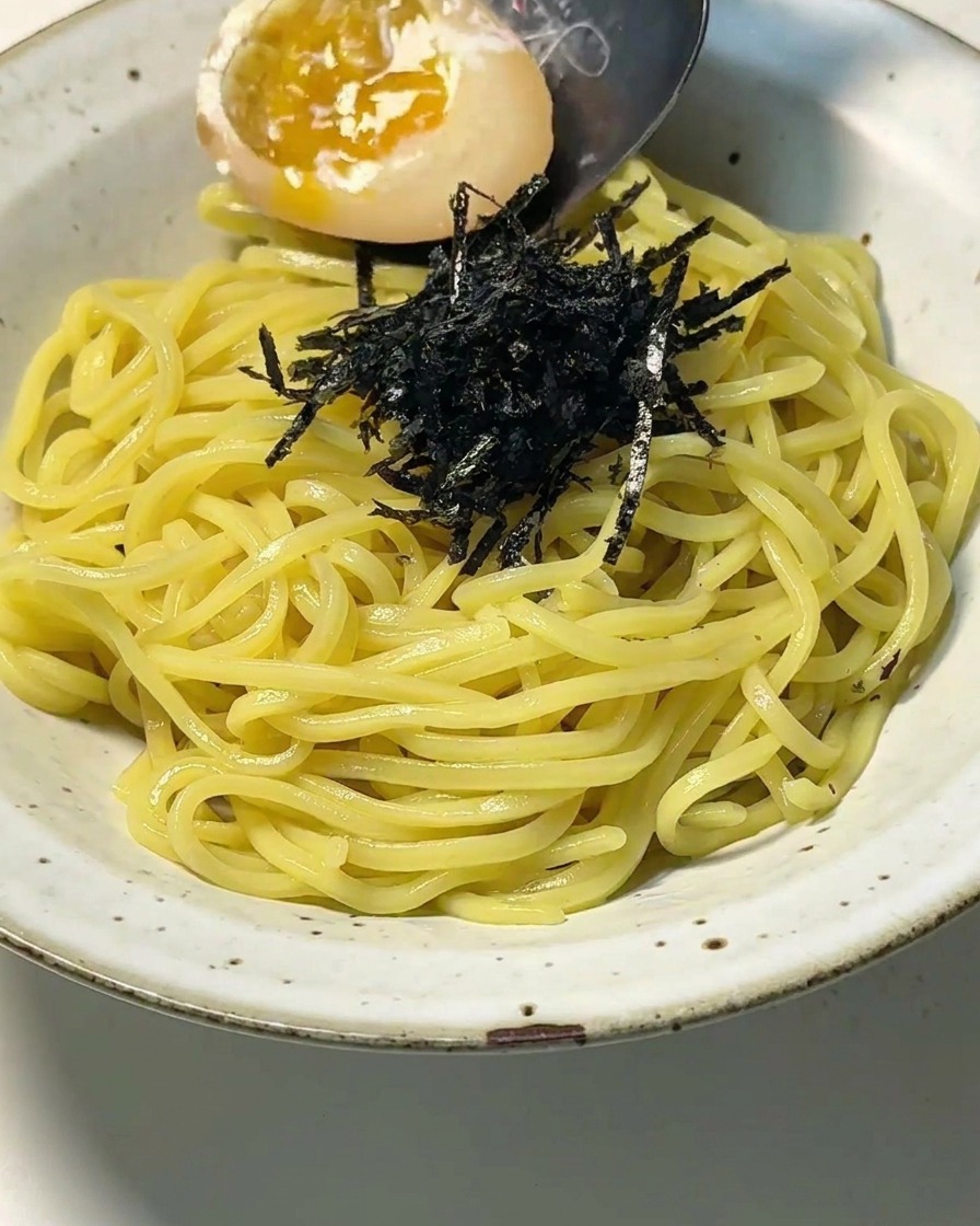 Primo piano di una ciotola con noodles e nori, mentre un cucchiaio adagia un uovo tagliato con tuorlo cremoso.