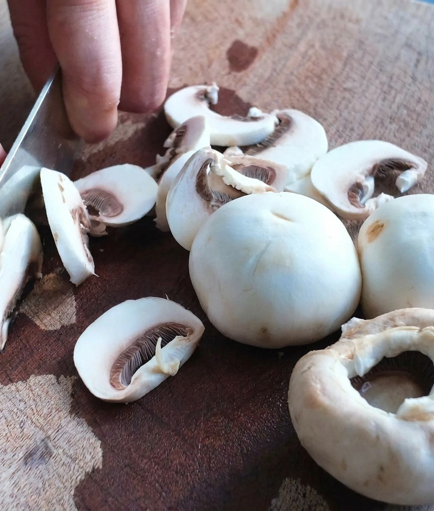 Primo piano di un coltello da chef che affetta funghi bianchi su un tagliere di legno testurizzato.