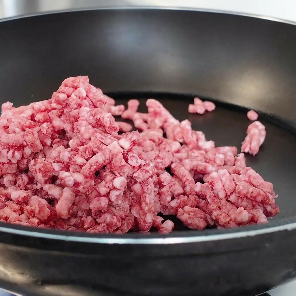 Carne macinata di manzo rosa che viene sgranata e saltata in una padella antiaderente nera.
