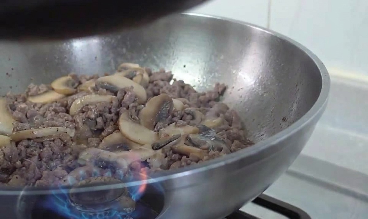 Un wok in acciaio inossidabile contenente un mix di carne rosolata e fette di funghi saltati su una fiamma a gas.