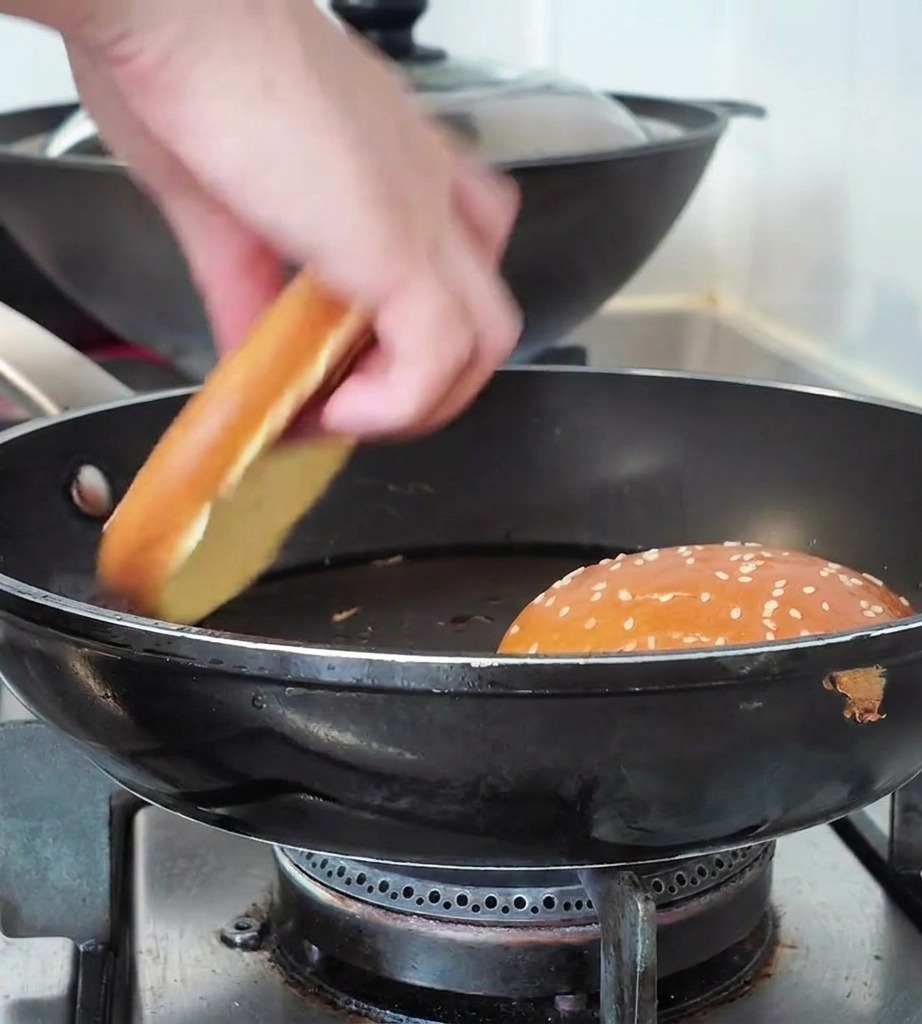 Una mano che posiziona metà di un panino al sesamo in una padella nera accanto a unaltra metà già in tostatura.