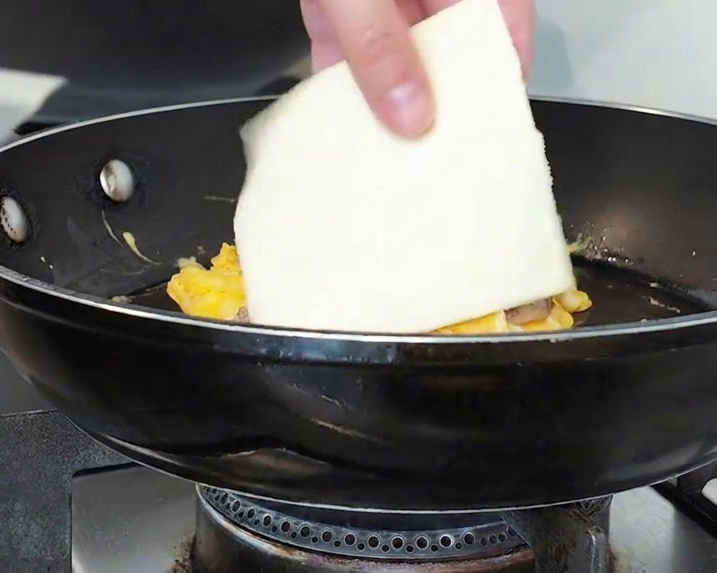 Una mano che mette una fetta quadrata di formaggio bianco sopra una montagna di carne e uova in padella.