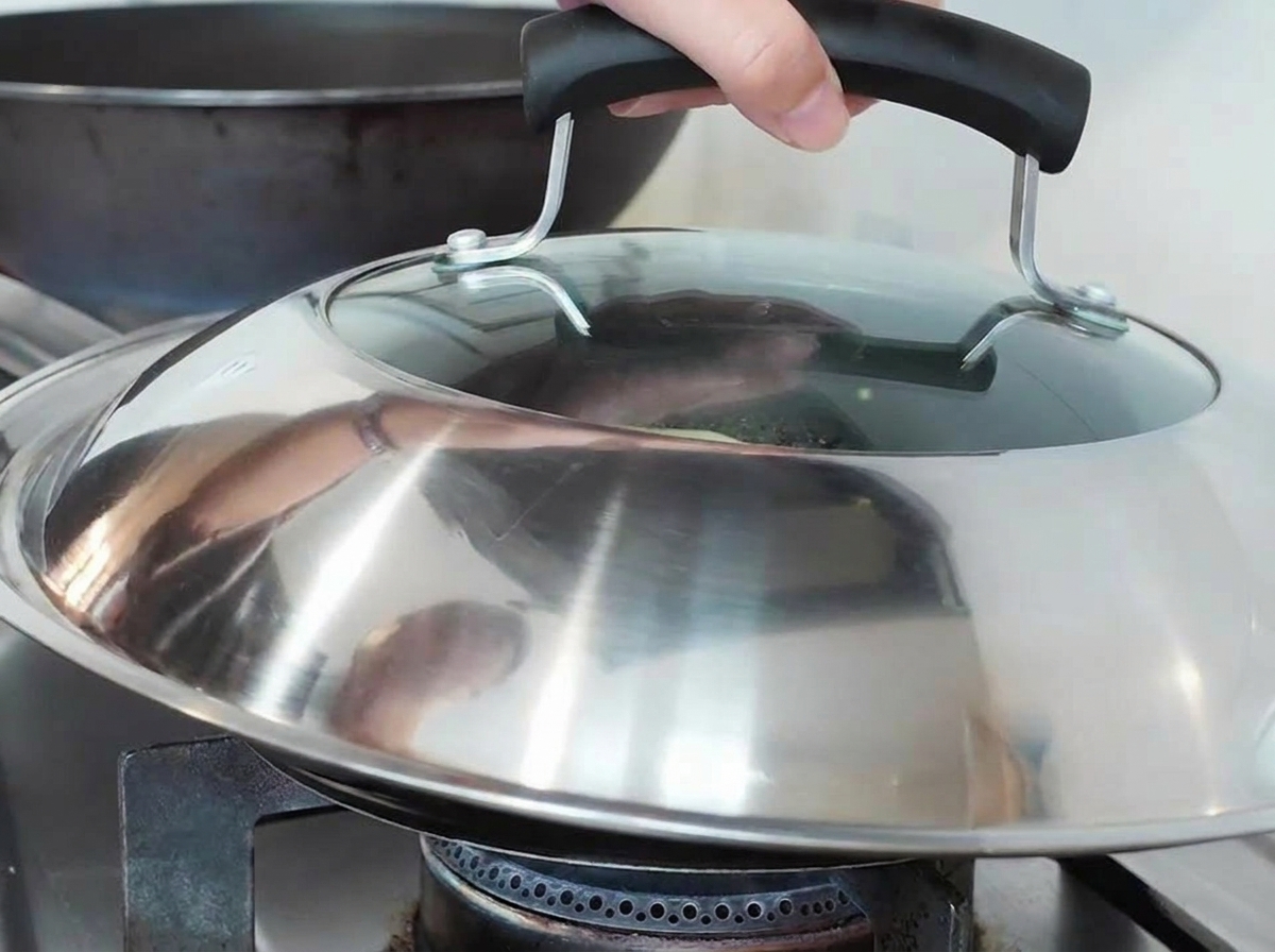 Una mano che mette un grande coperchio di metallo e vetro sopra una padella su un fornello a gas.