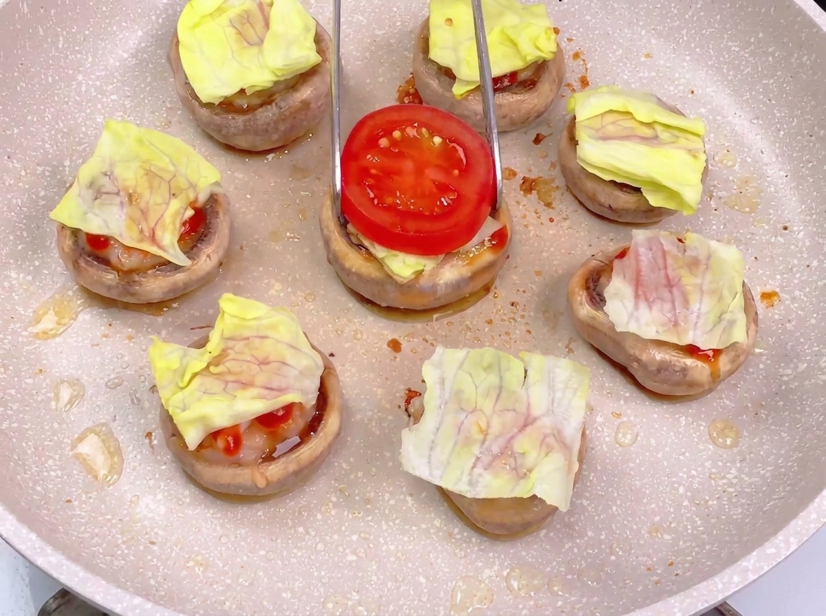 Delle pinze che posizionano una fetta di pomodoro rosso sopra un mini burger già stratificato con lattuga e salse.