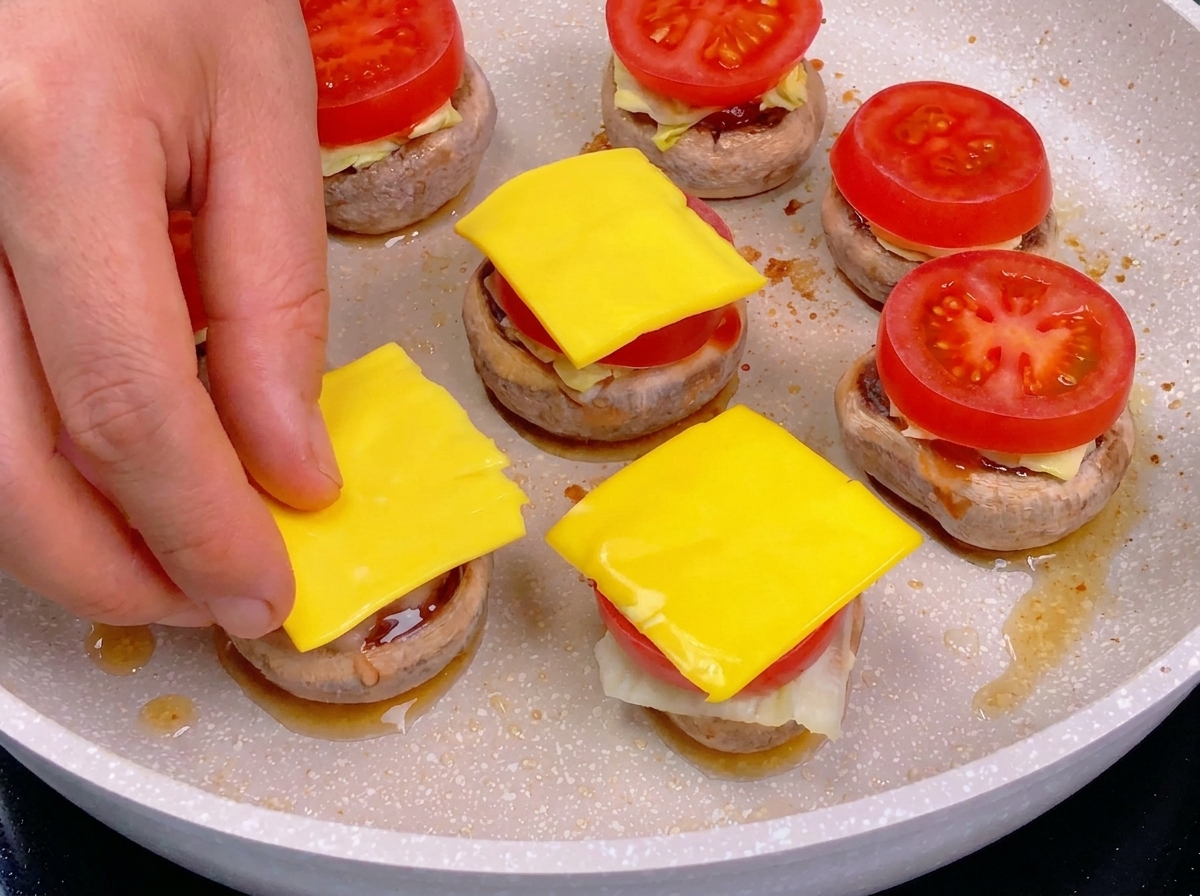 Una mano che mette un quadratino di formaggio giallo sopra una fetta di pomodoro in un mini burger di funghi.