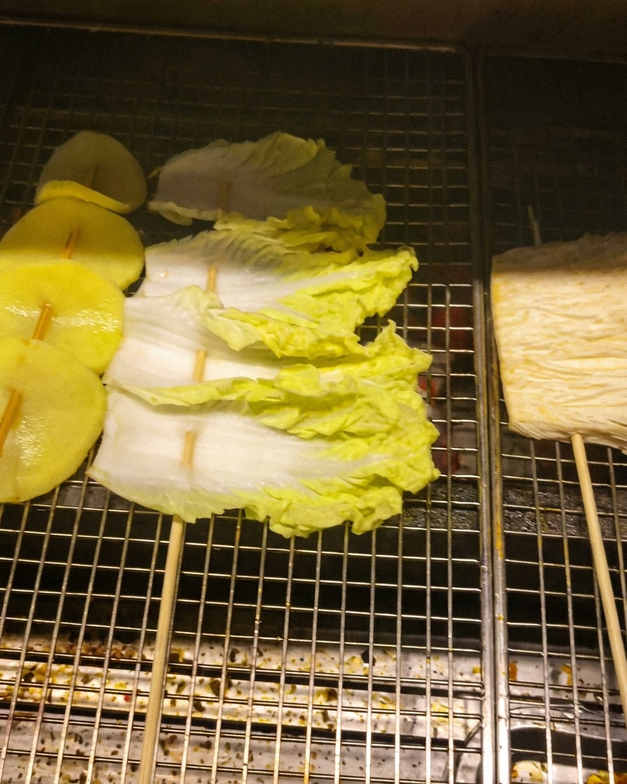 Fette di patata, foglie di cavolo e funghi enoki infilzati in spiedini sulla griglia metallica.