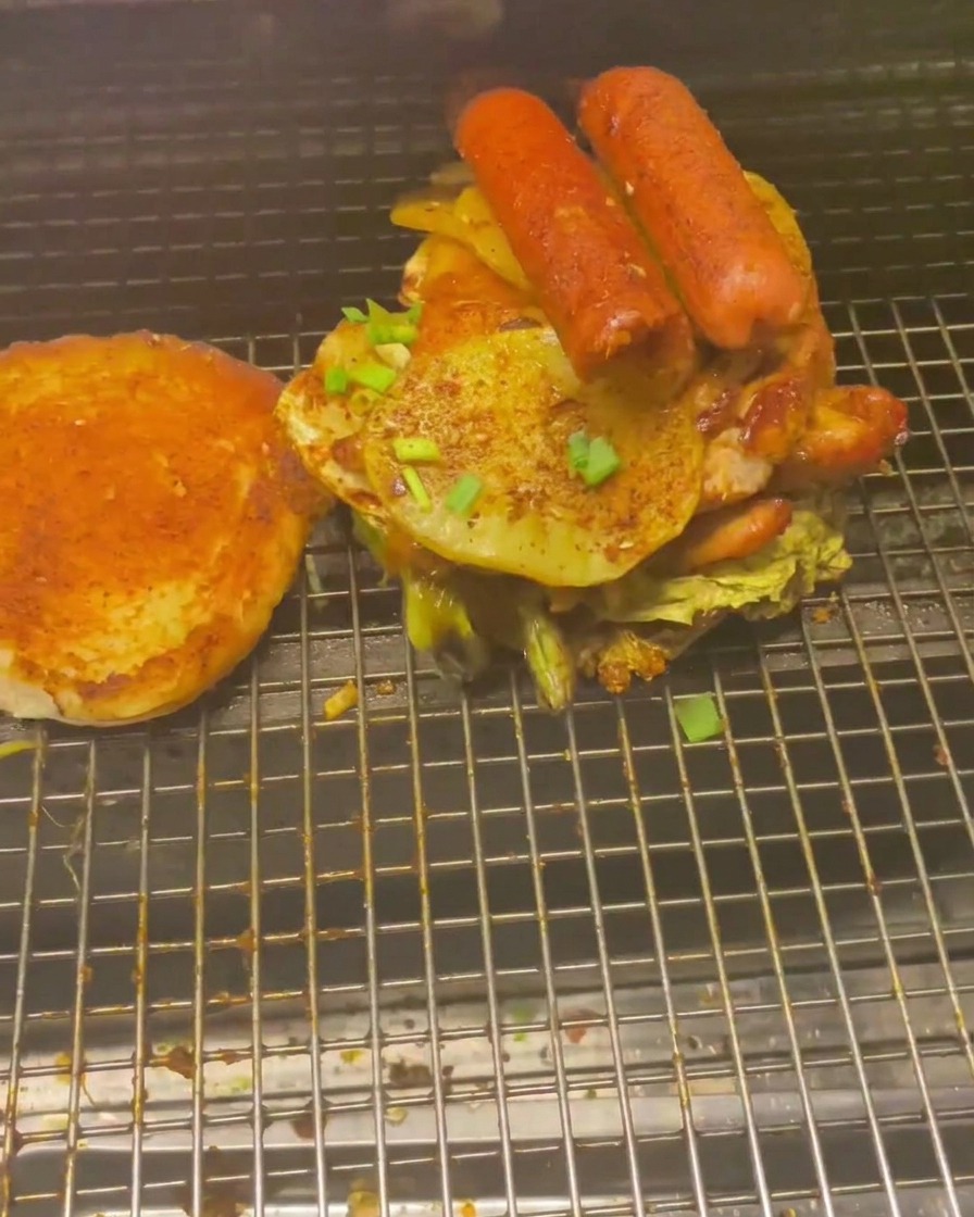 Pila alta di carni e verdure, inclusi würstel e fette di patata, sopra un panino.
