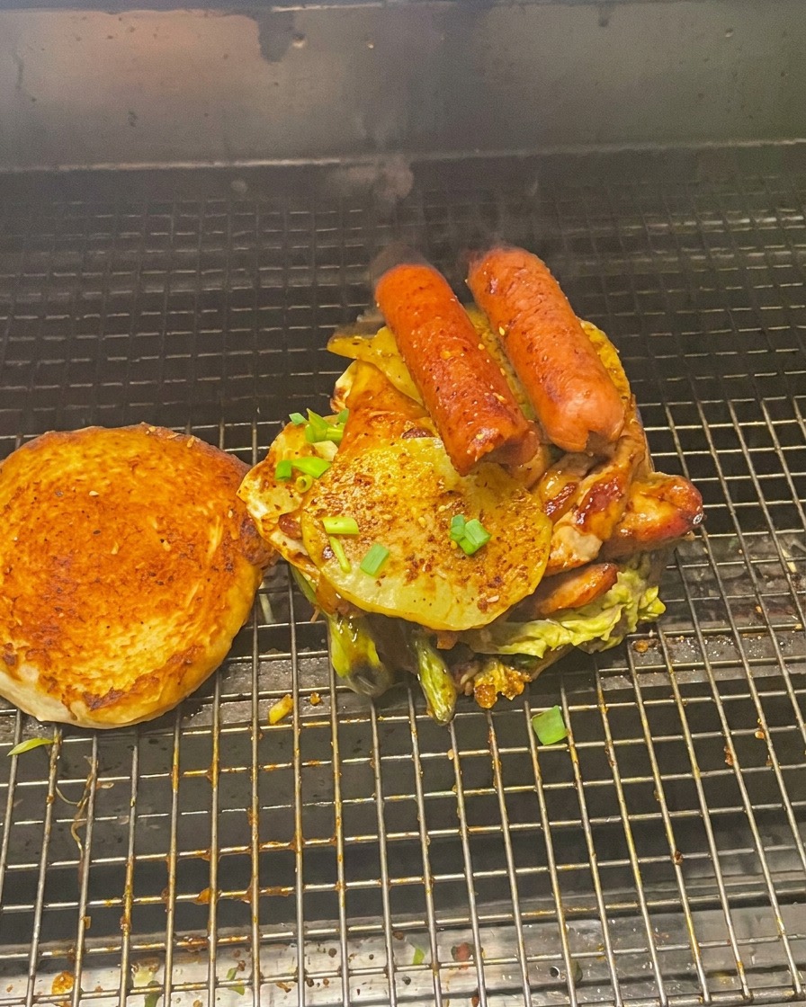 Imponente pila di carne e verdure sulla base del panino, con la parte superiore tostata accanto sulla griglia.