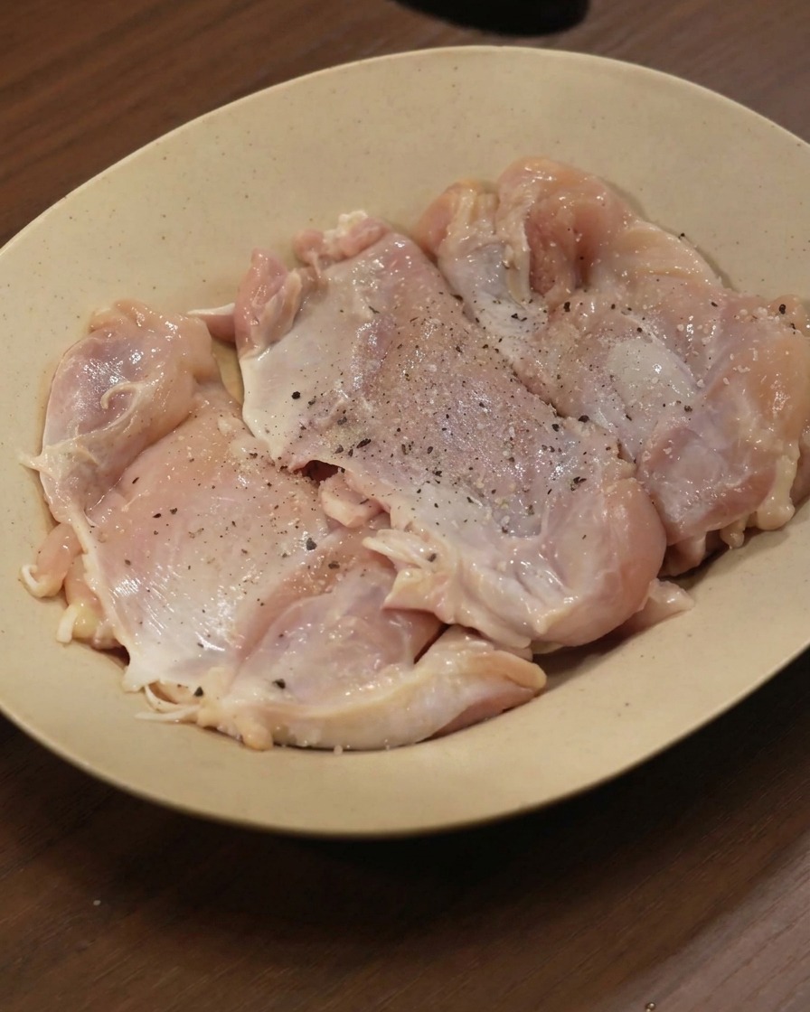 Sovracosce di pollo crudo disposte su un piatto beige, cosparse di pepe nero e varie spezie in polvere.
