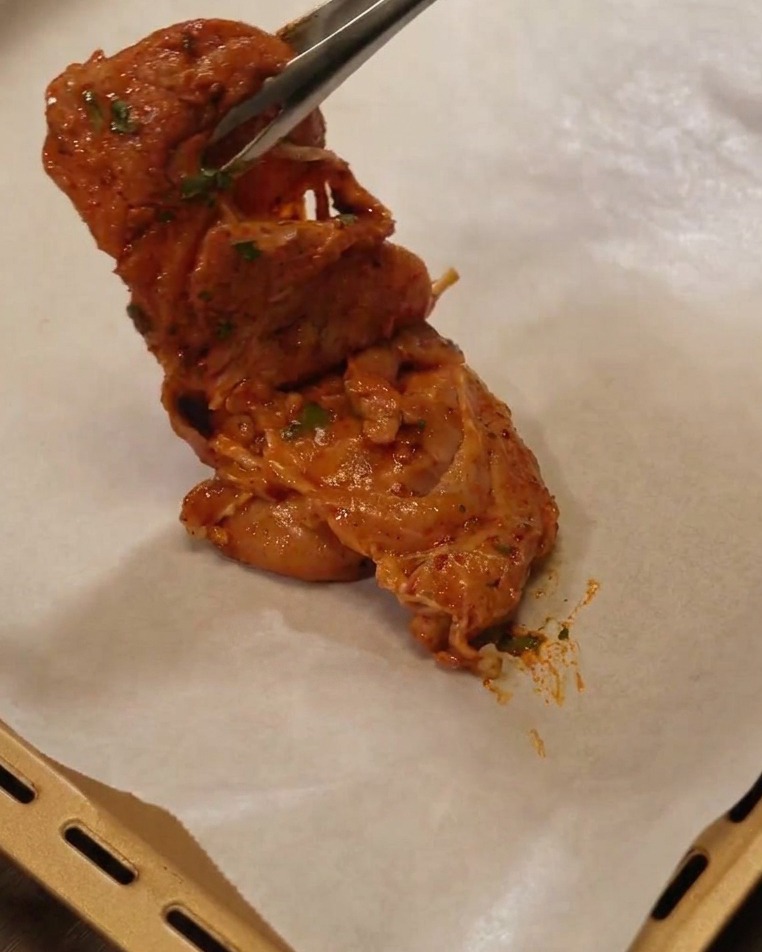 Pinze metalliche che sollevano una sovracoscia di pollo marinata posizionandola su una teglia foderata con carta forno.