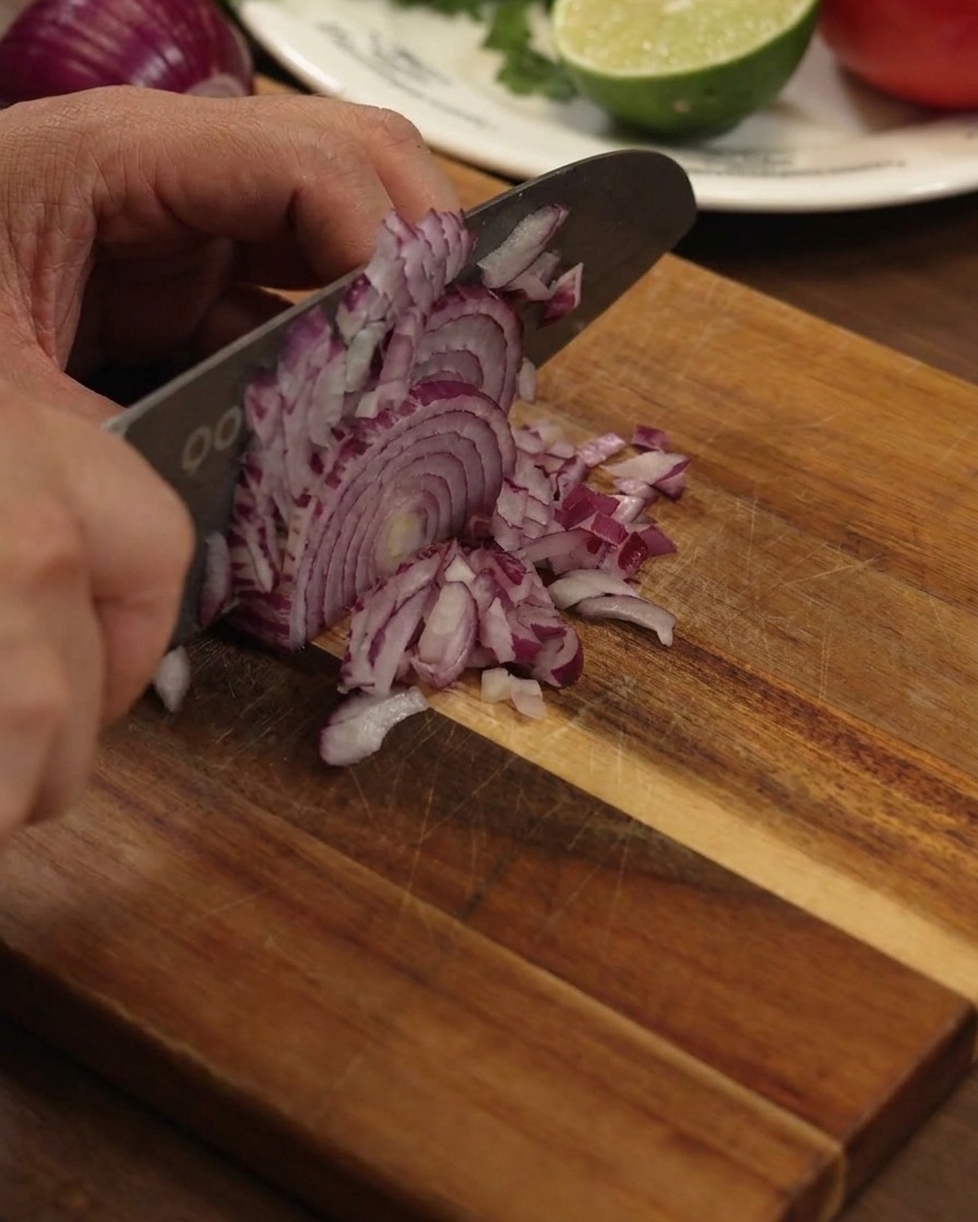 Una mano che usa un grande coltello da cucina per tagliare finemente una cipolla rossa su un tagliere di legno.