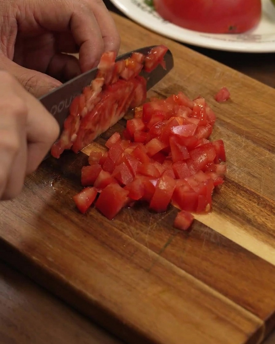 Taglio di un pomodoro rosso maturo in piccoli cubetti regolari su un tagliere di legno.