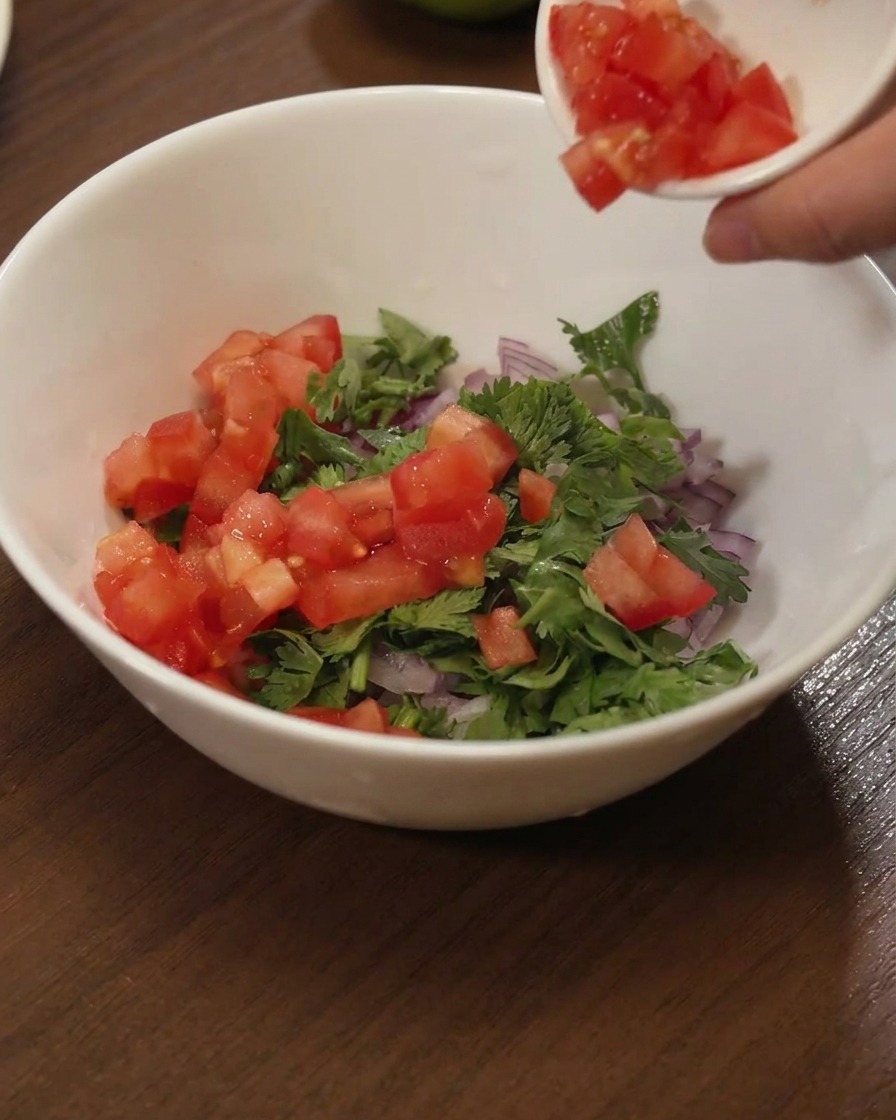 Pomodori rossi a cubetti versati da un piattino in una ciotola bianca con coriandolo e cipolle rosse.
