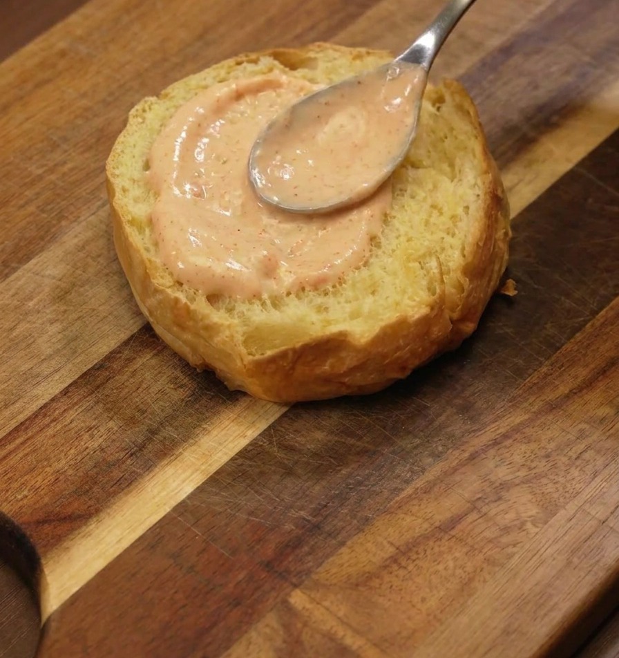 Un cucchiaio dargento che spalma uno strato di salsa burger arancione sulla parte tostata di un panino brioche.