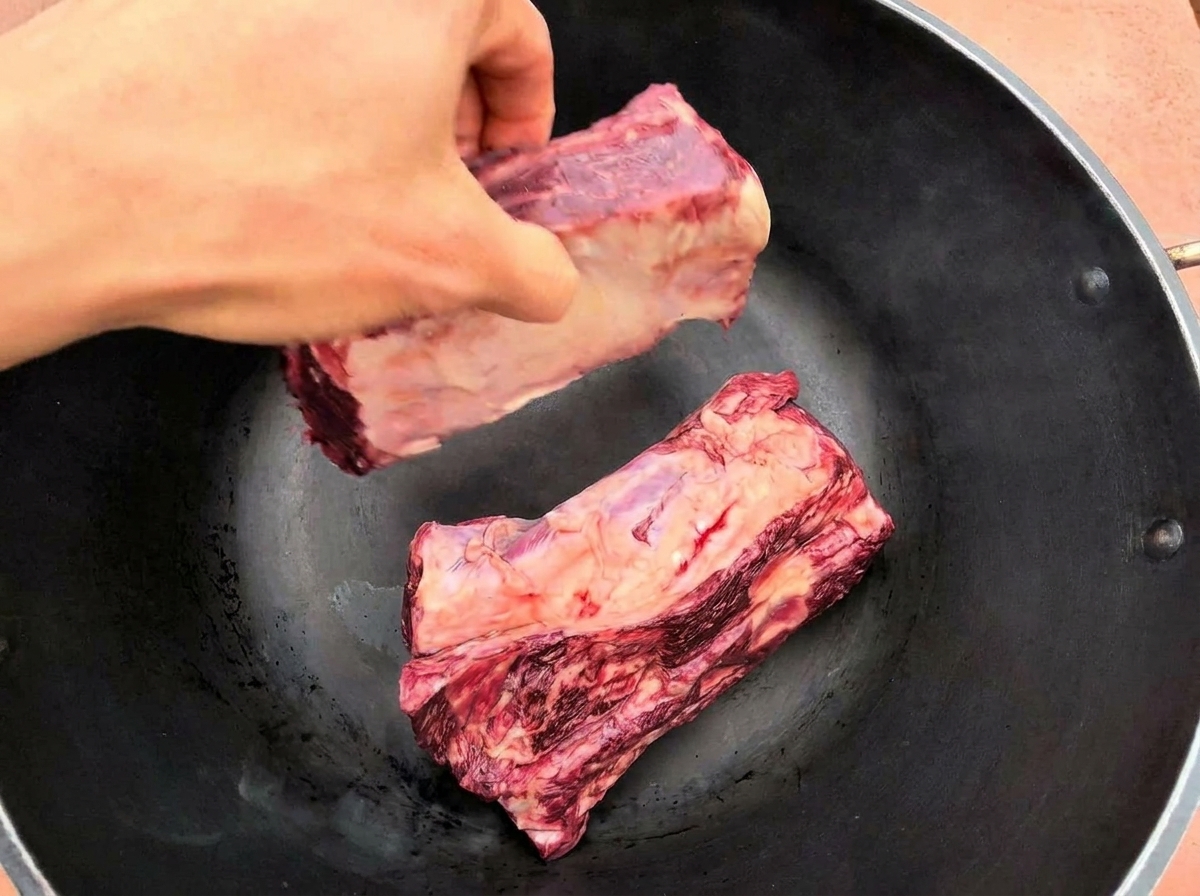 Una mano adagia due grandi costine di manzo crude in una padella nera ben calda.