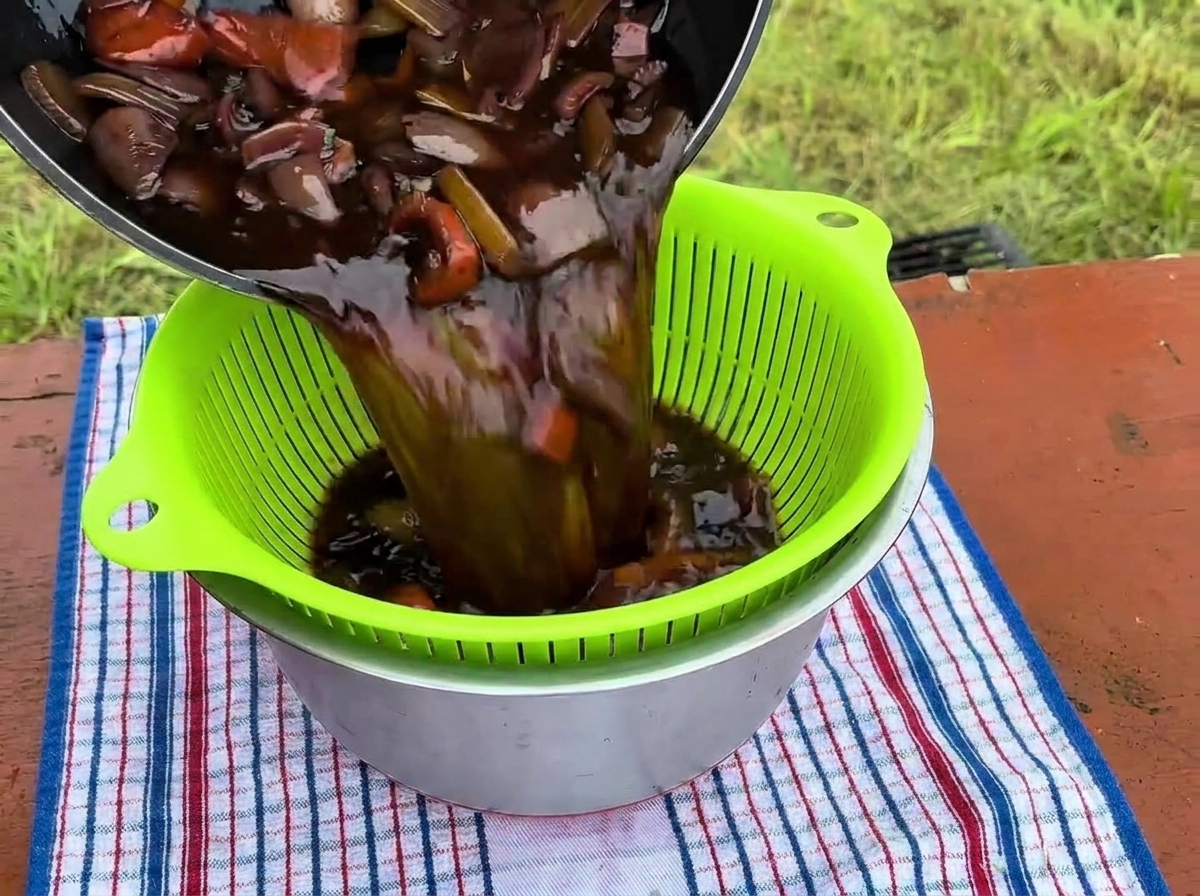Un ricco e scuro liquido di brasatura insieme alle verdure cotte viene versato da una pentola in un colino verde brillante posizionato sopra una ciotola in acciaio inossidabile.