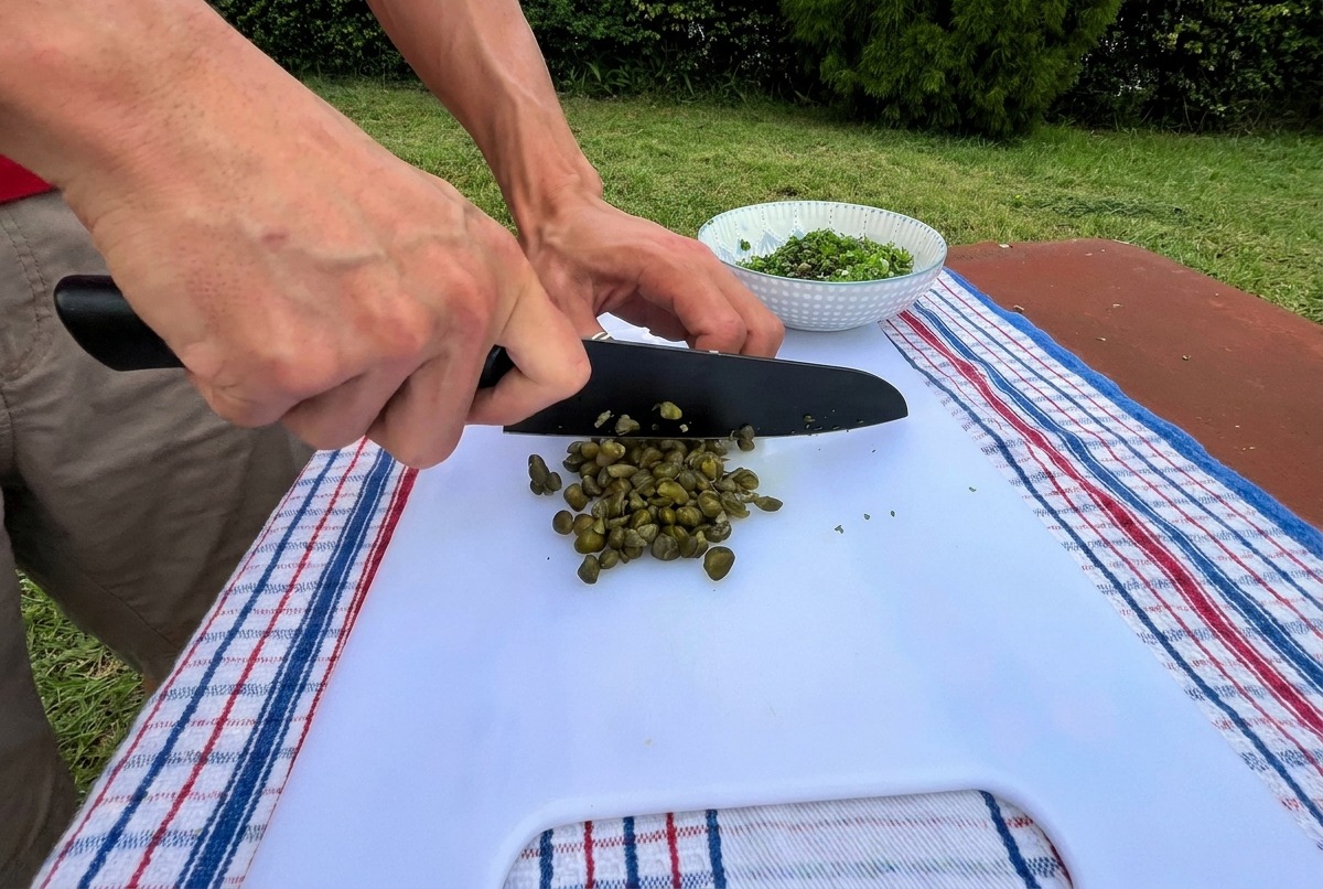 Primo piano di una persona che usa un coltello da chef nero per tritare finemente dei capperi verdi su un tagliere bianco allaperto.