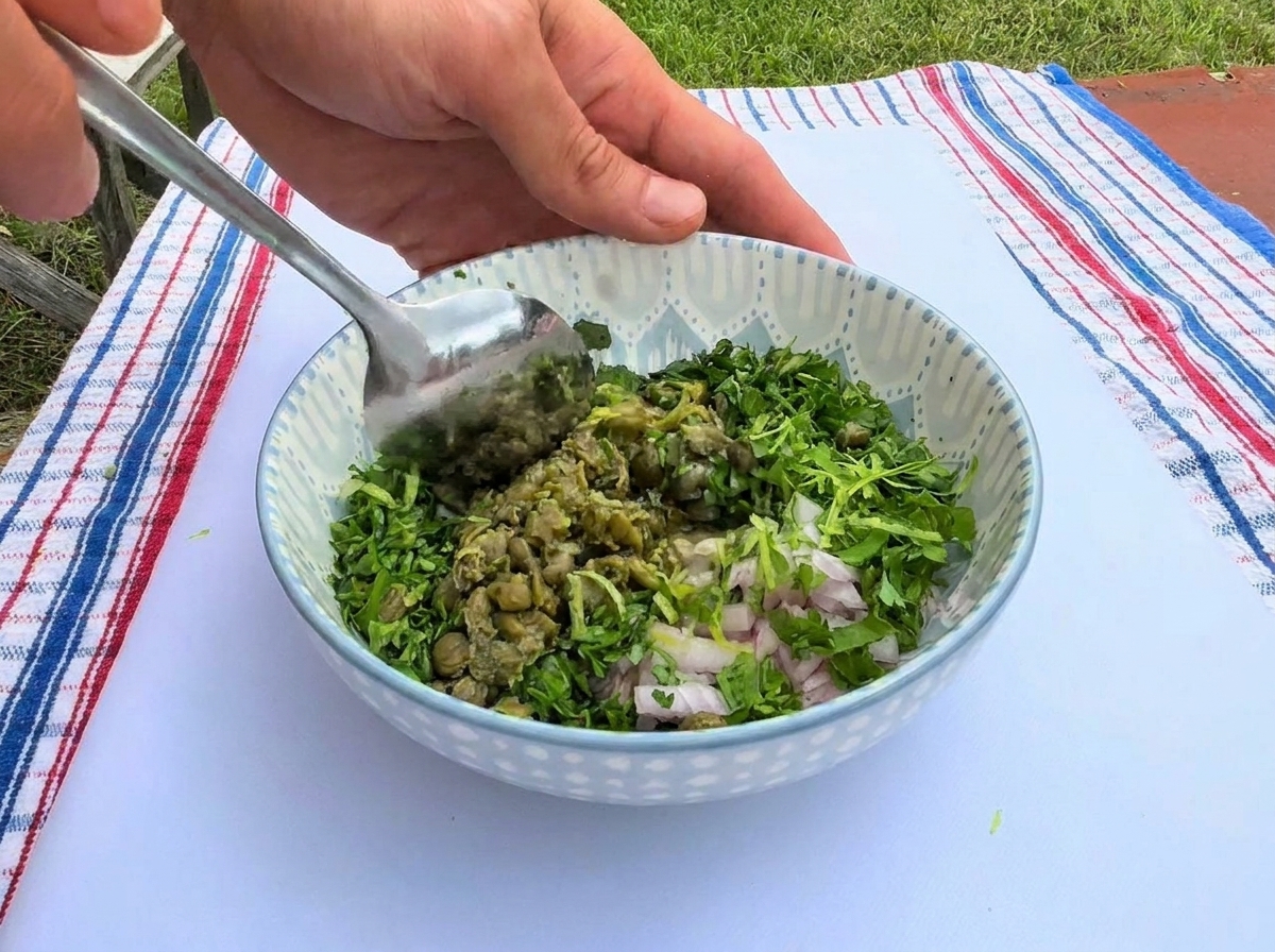 Mescolando un vibrante composto di erbe verdi tritate, capperi e scalogno sminuzzato con un cucchiaio di metallo allinterno di una ciotola di ceramica decorata.