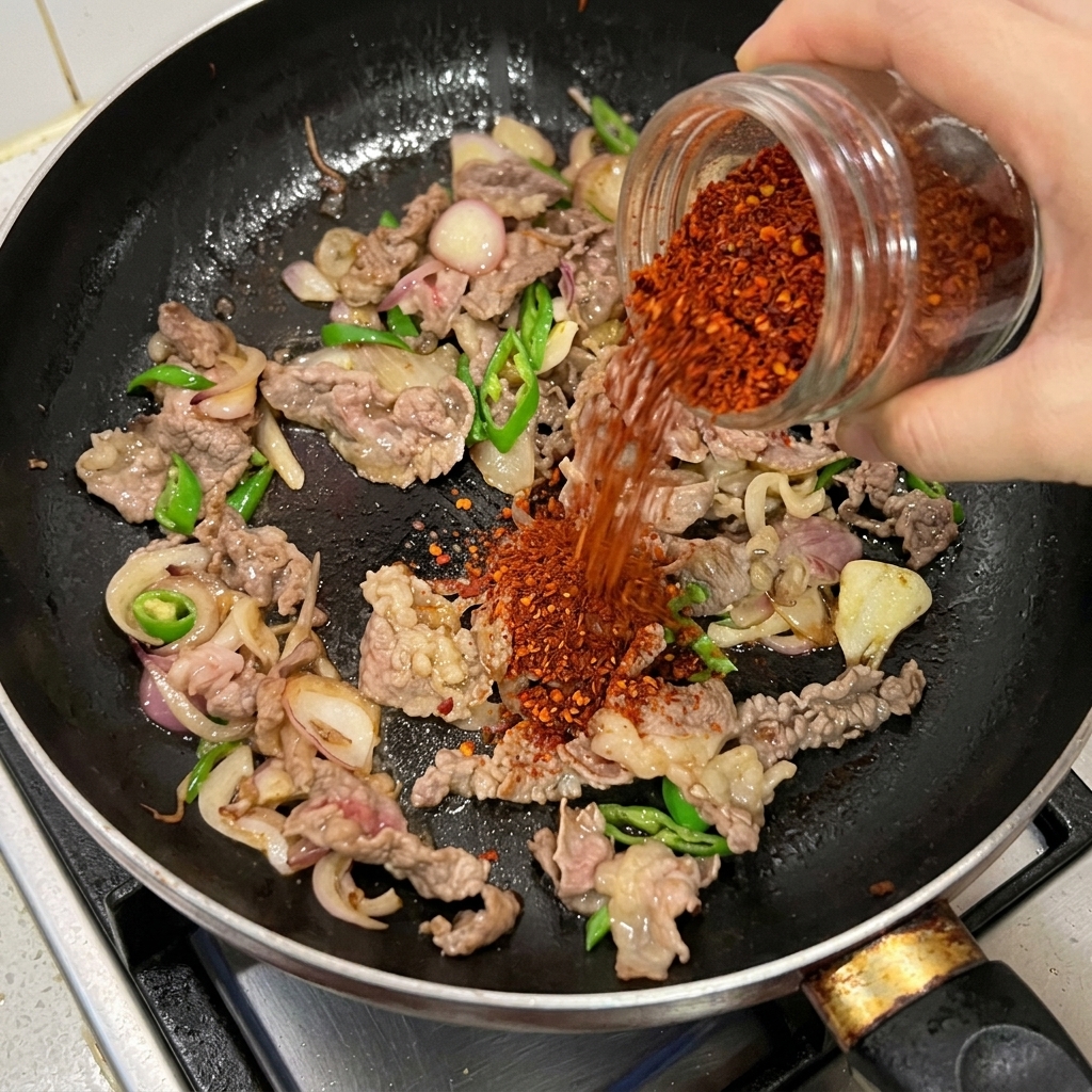 Peperoncino rosso in polvere versato da un barattolo di vetro su carne, cipolle e peperoncini verdi in padella.